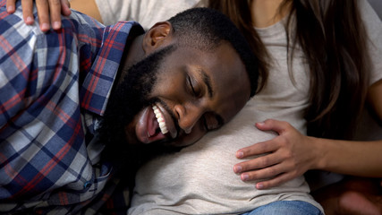 Cheerful Afro-American man listening to baby in belly, happy expectation, family