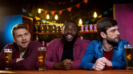 African-american man smiling watching tv in bar, caucasian friends upset