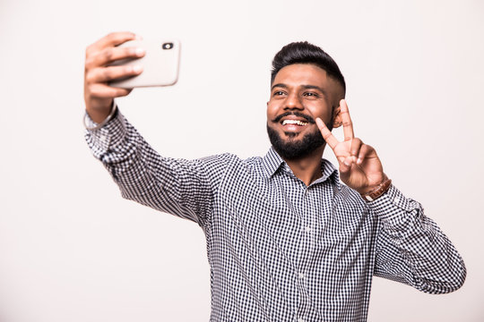 Young indian young man with beard and mustache taking selfie using smartphone make peace gesture, isolated over white background