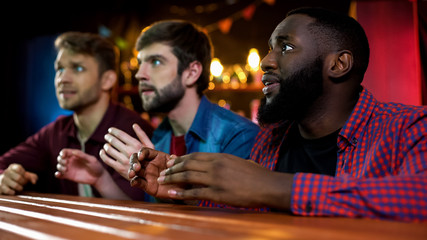 Three multiethnic friends watching song contest on tv in bar, ready for applause
