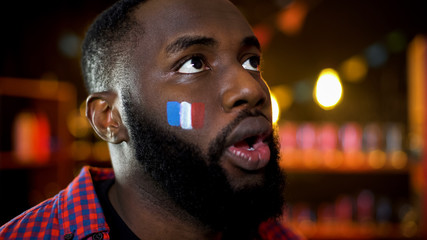Shocked african-american man with painted french flag on cheek watching news