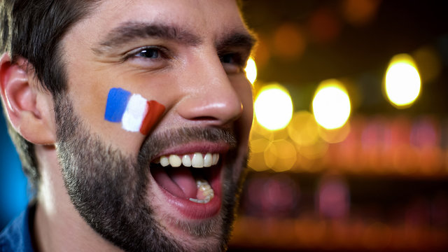 Happy handsome bearded french fan with painted flag on cheek celebrating goal - Powered by Adobe