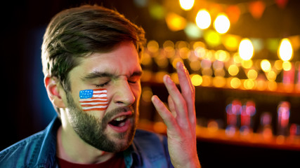Fototapeta premium Anxious football fan with american flag on cheek displeased with game result