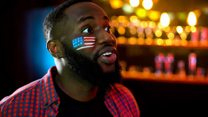 Anxious black man with usa flag on cheek watching sport competition on tv in bar