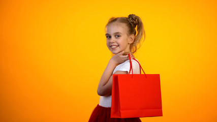 Fototapeta premium Cute little girl with red shopping bag smiling and to camera, shopping day