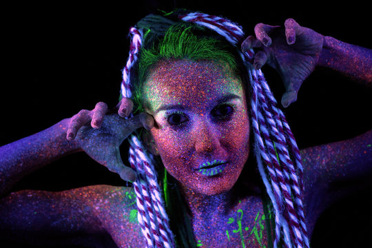 The girl aliens looks to the camera. shrugs her shoulders, ultraviolet make-up.  She holds her hands near her face, next to her ears.