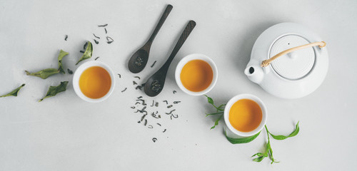 Tea concept with three white cups of tea and teapot surrounded with fresh green tea leaves and dry leaves,  top view composition on concrete background with copy space. Long banner.