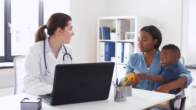 medicine, healthcare and pediatry concept - african american mother with baby son and caucasian doctor with laptop computer at clinic