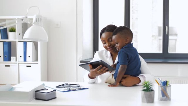 medicine, technology and pediatry concept - happy african american female doctor or pediatrician showing tablet computer to baby boy patient at clinic