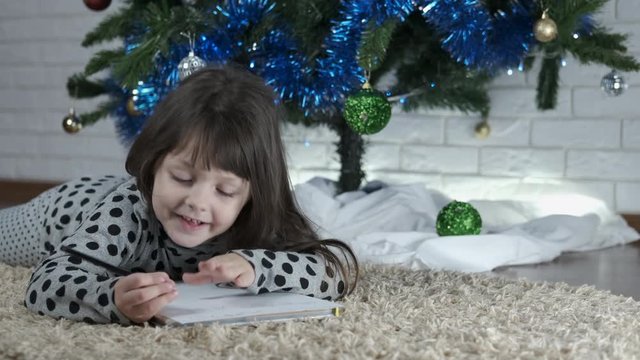 Thinking about Christmas. Cute little girl writes a letter under the Christmas tree. Write a Christmas letter.