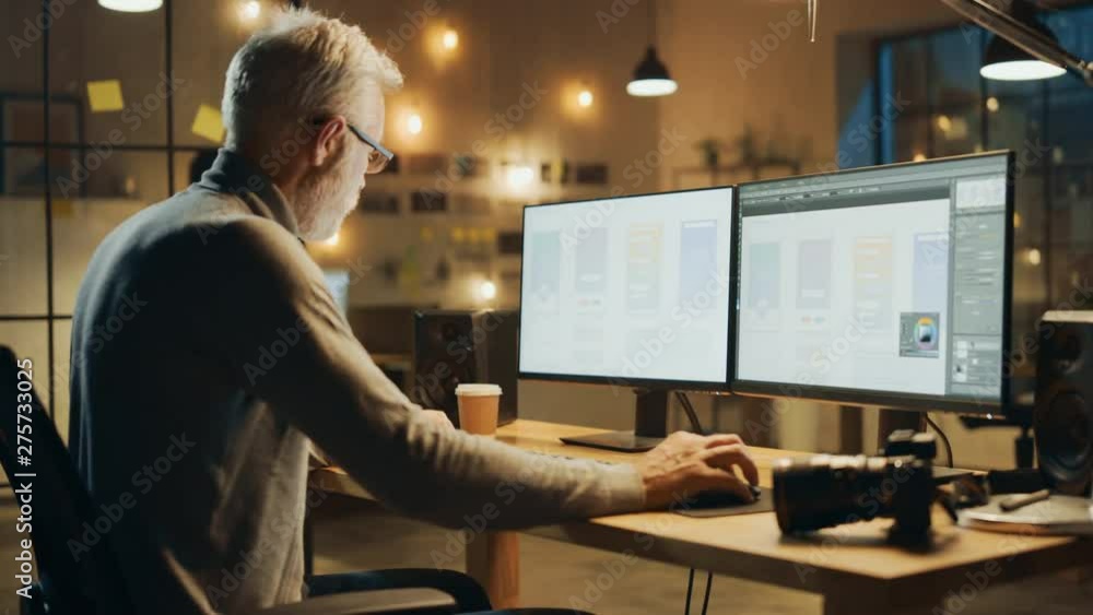 Poster creative middle aged mobile software designer sitting at his desk uses desktop computer with two scr