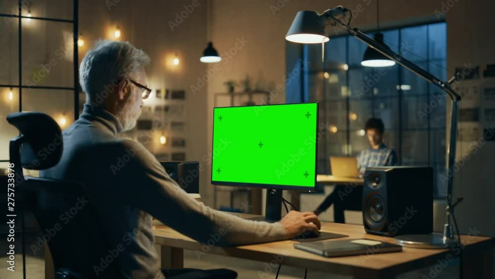 Poster Creative Middle Aged Designer Sitting at His Desk Uses Desktop Computer with Green Mock-up Screen. In the Evening Two Creative Employees Working on Computers in the Office with Cityscape Window View