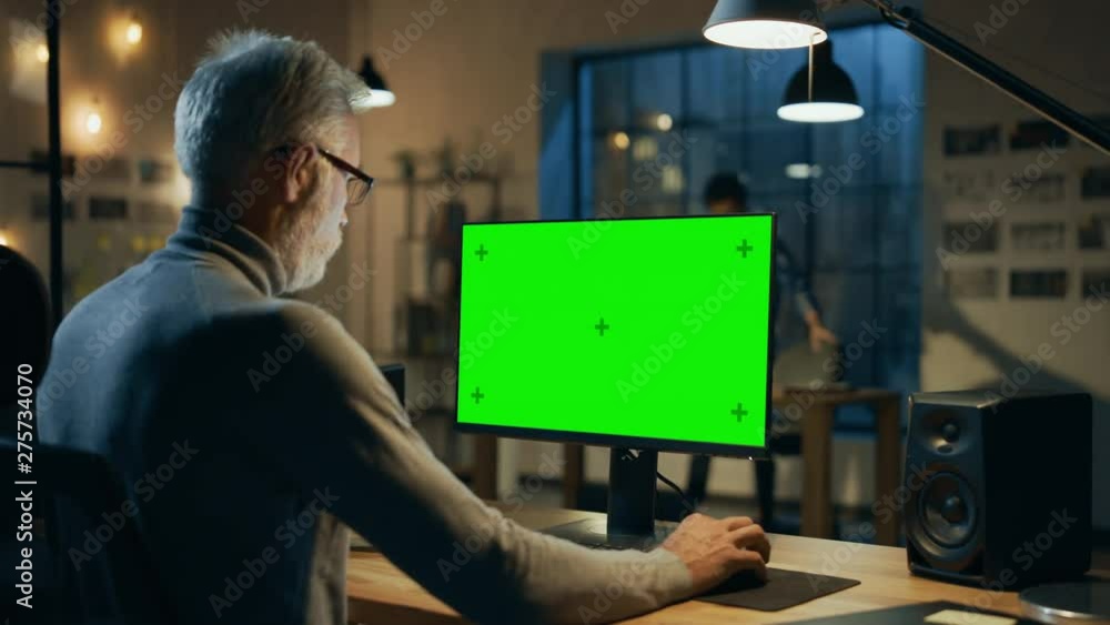 Poster creative middle aged designer sitting at his desk uses desktop computer with green mock-up screen. i