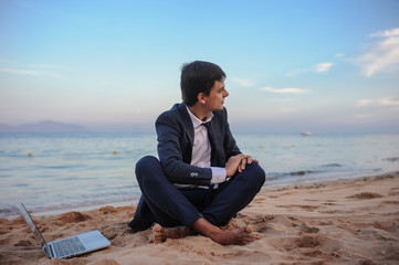 lateral view of a young man in suit with laptop sitting on the beach working on the seaside