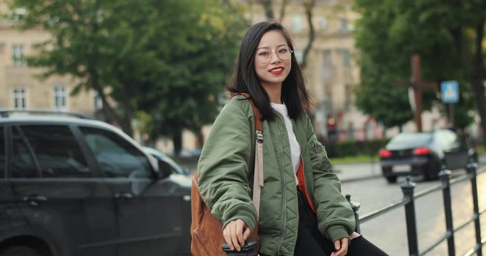 Portrait shot of the stylish Asian young girl tourist in glasses standing at the street and posing to the camera.