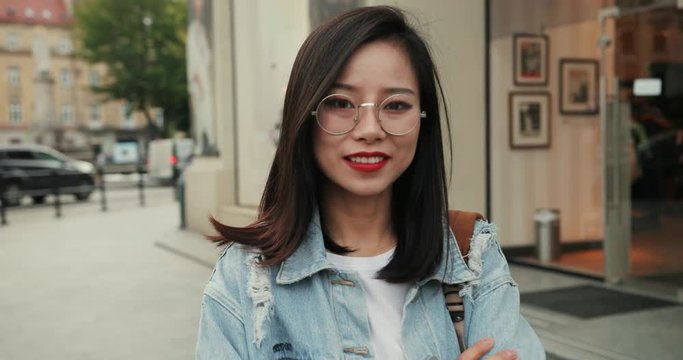 Portrait of the attractive young Asian woman with black hair and in glasses posing to the camera and smiling outdoor in the city. Close up.