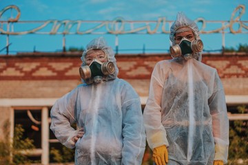 Obraz na płótnie Canvas Two scientists put on a respirator and a radiation protective suit. Stern look into the camera from the abandoned territory. Care about ecology in bright saturated photos.