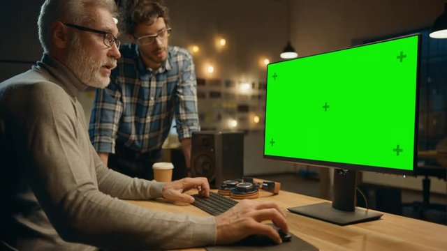 In the Evening Creative Middle Aged Man Works on a Desktop Computer Green Mock-up Screen, Has Conversation with Younger Specialist who Uses Tablet Computer