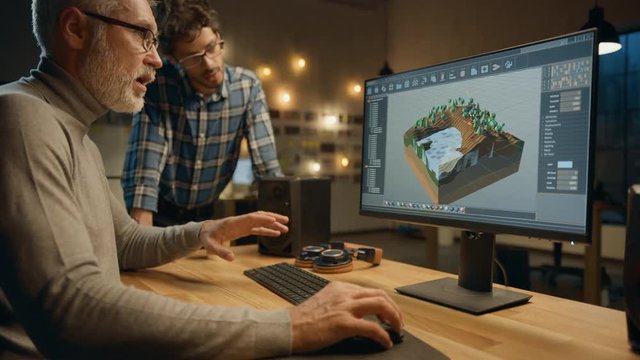 In the Evening Creative Middle Aged Video Game Developer Works on a Desktop Computer with Screen 3D Videogame Level Design, Has Conversation with Younger Specialist who Uses Tablet Computer