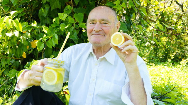 Very old man is drinking a homemade limonade with a yellow reusable boba straw. Elderly gentleman drinks a lemon cocktail on green vegetation background.The concept of a healthy lifestyle and natural 