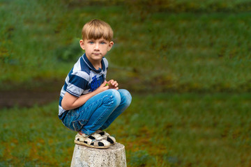 A boy in blue jeans squats in the park looking around.