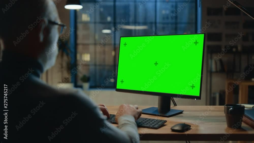 Poster Over the Shoulder: Confident Middle Aged Man Sitting at His Desk Using Desktop Computer with Mock-up Green Screen. Evening in the Stylish Office Studio with City Window View