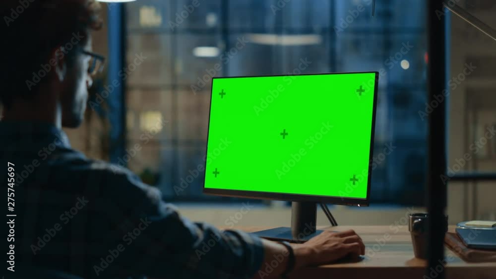 Poster Over the Shoulder: Creative Young Man Sitting at His Desk Using Desktop Computer with Mock-up Green Screen. Evening in the Stylish Office Studio with City Window View