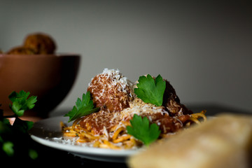 A close-up shot of a plate of rustic spaghetti and meatballs with a bowl of meatballs, parmesean cheese, and fresh parsley.