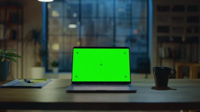 Mock-up Green Screen Laptop Standing on the Desk in the Modern Creative Office. In the Background Warm Evening Lighting and Open Space Studio with City Window View. Zoom in Shot