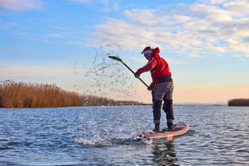 Man paddle (floating) on Stand up paddle board (SUP ) in Santa Claus costume in calm of winter lake...