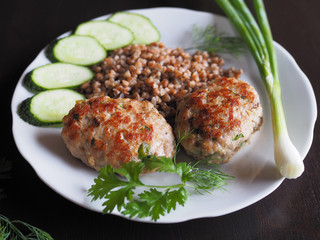 Appetizing homemade chopped steak with buckwheat to eat with herbs and vegetables. Cutlets with vegetables and garnish are in a white plate on a dark table. 