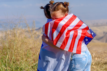 Two cute girls with American and Israel flags. Little children holding Israeli and USA flags hugging on meadow with beautiful landscape in background.