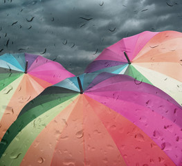 Rainbow umbrellas on dramatic sky background under rain, view through window glass with rain drops