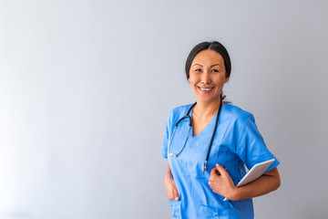 Portrait of confident young Asian female doctor. Portrait of a good looking young nurse with tablet computer in a grey background. Doctor with stethoscope around his neck looking at the camera