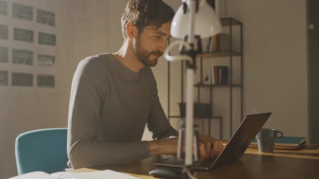 Professional Creative Man Sitting at His Desk in Home Office Studio Working on a Laptop Drinks Beverage from the Cup, Writes Down Ideas in Notepad. Bright Modern Room. 360 Degree Tracking Arc Shot