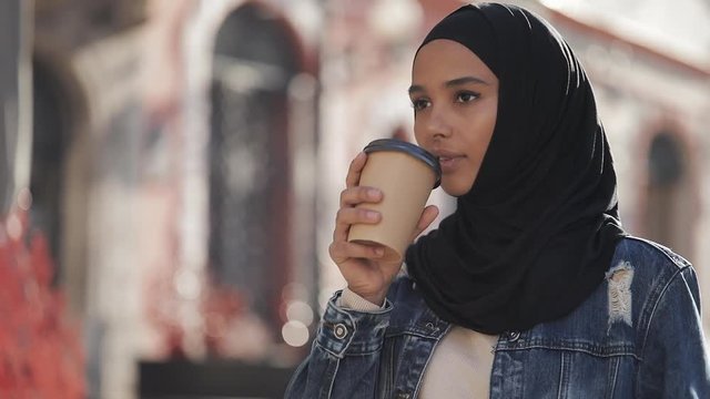 Beautiful young Muslim woman an attractive appearance drinking coffee and walking down the city street.