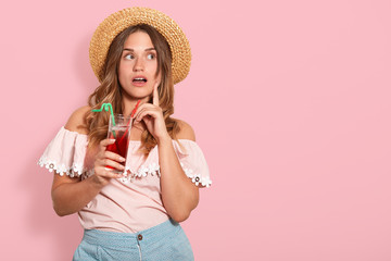 Close up studio photo portrait of thoughtful pondering female, attractive lady holding tasty beverage in hand, wearing straw sunhat and shirt, model posing isolated rose background. People concept.