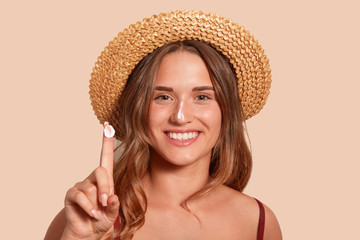Young woman applaying sunblock on her face, and showinig her finger with sunscreen, model posing isolated over beige background, standing against studio wall with topothy smile, dressed swimming suit.