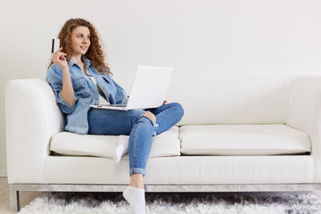 Portrait of beautiful positive lady looking aside, raising hand with credit card, making plans for online shopping, having laptop on legs, wearing jeans and jacket, having pleasant facial expression.