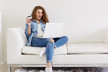 Fototapeta na wymiar Charismatic attractive curly haired woman opening her mouth widely with satisfaction, looking at laptop screen attentively, holding credit card in hand, being concentrated on shopping. Rest concept.