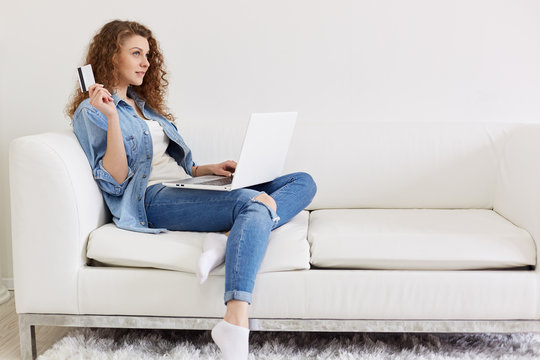 Portrait of thoughtful magnetic woman sitting with her laptop on sofa, holding credit card in one hand, thinking about purchases online, like spending money, wearing casual jeans clothes. Shopping.