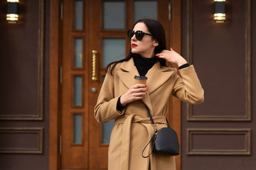 Image of stylish beautiful female standing outside, wearing beige coat, black shirt, fashionable sunglasses, touches her hair, having paper cup of coffee or tea in other hand, looking aside.