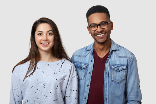 Horizontal shot of happy dark skinned young man and European woman, standing with positive expressions, dressed in casual clothes, posing smiling indoor isolated over white studio background.