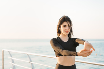Pretty young brunette woman in a black jumpsuit stretches on a pier at sunrise before exercising.