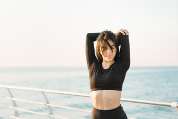 Pretty young brunette woman in a black jumpsuit stretches on a pier at sunrise before exercising.