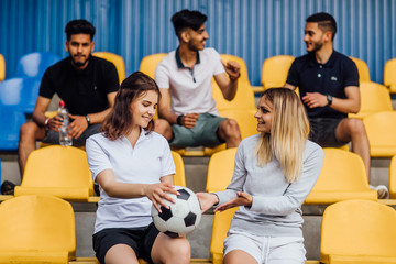 Friends in sports clothing smiling while sitting on the stadium, outdoors. Talking after play.