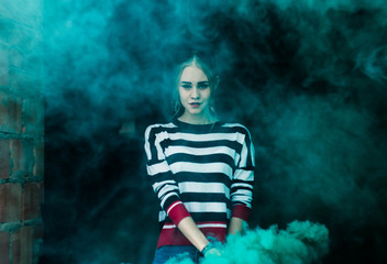 Fototapeta na wymiar Blonde in a striped jacket on the background of a brick building and trees with a green smoke bomb