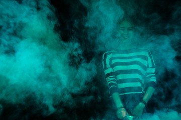 Blonde in a striped jacket on the background of a brick building and trees with a green smoke bomb - Powered by Adobe