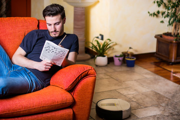 Young man sitting doing a crossword puzzle looking thoughtfully at a magazine, with his pencil to...