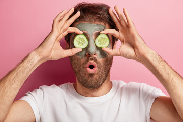 Unshaven man keeps face in good condition, covers eyes with two slices of cucumber, has clay mask on skin, cares about beauty, has rejuvenation treatments at home, stands indoor in bathroom.